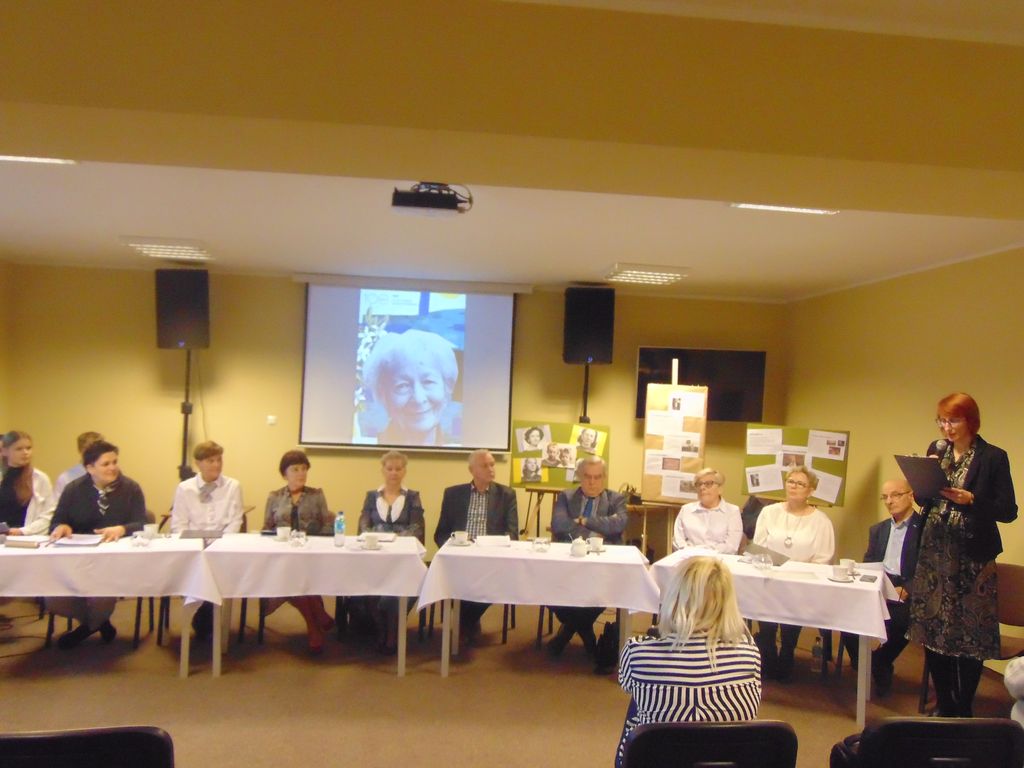 Uczestnicy spotkania siedzą w sali. Jedna osoba stoi i czyta przy mikrofonie.